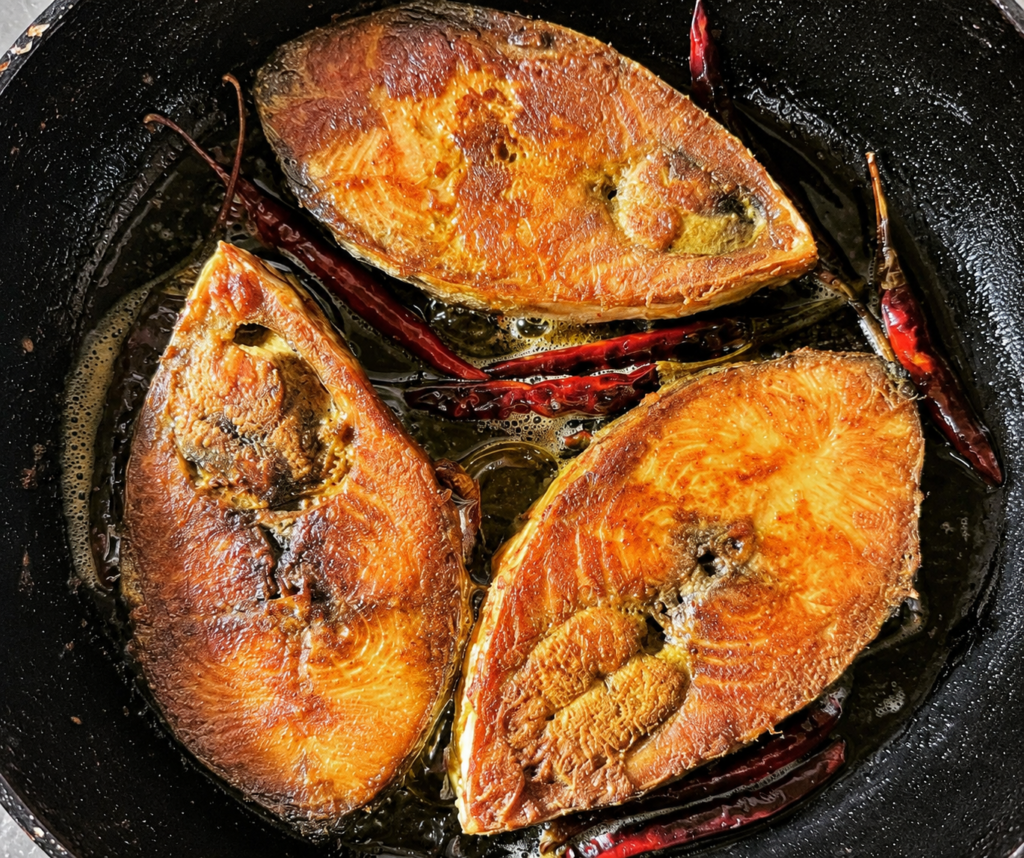Three golden pieces of hilsa fish frying with dried red chillies in a traditional black pan.