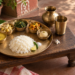 A large kansa thali with rice and five small bowls of Bengali vegetarian dishes, arranged on a weathered wooden jolchoki beside a kansa glass and ghot.